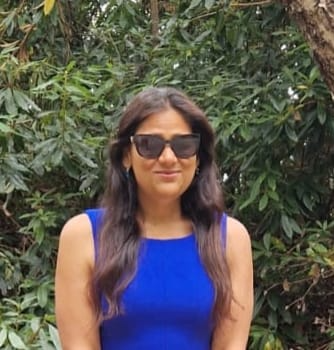 A dark-haired woman in her mid-thirties with olive toned skin and mid length wavy locks cascading down either side of her shoulders. She is wearing a sleeveless sapphire blue dress and sunglasses, affecting a soft smile while standing in front of lush greenery.