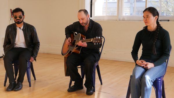 Three actors sitting on stage, from left to right Shashank, Chris, who is playing a guitar and Jasmine.