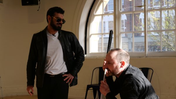 Shashank has his arm on his hip and talks to Chris who holds his cane and crouches down towards the floor.