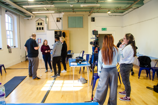 Tam is speaking to 3 actors in a rehearsal room. Next to them is a table and camera equipment with people filming.